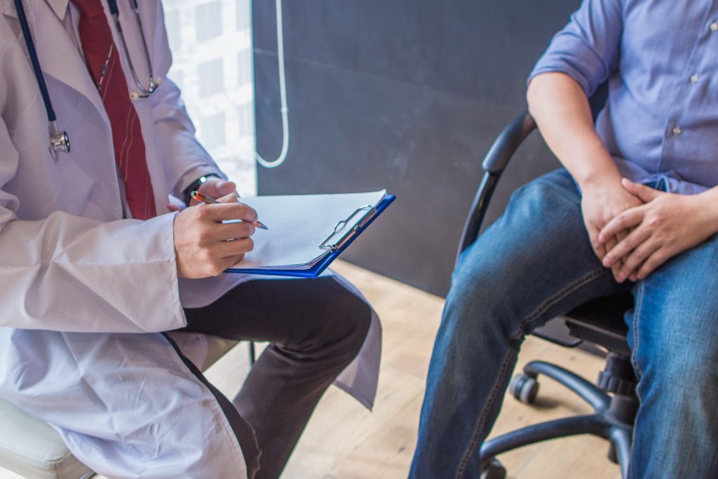 Doctor writing notes on clipboard while patient sits in chair, doctor holding pen and talking to patient