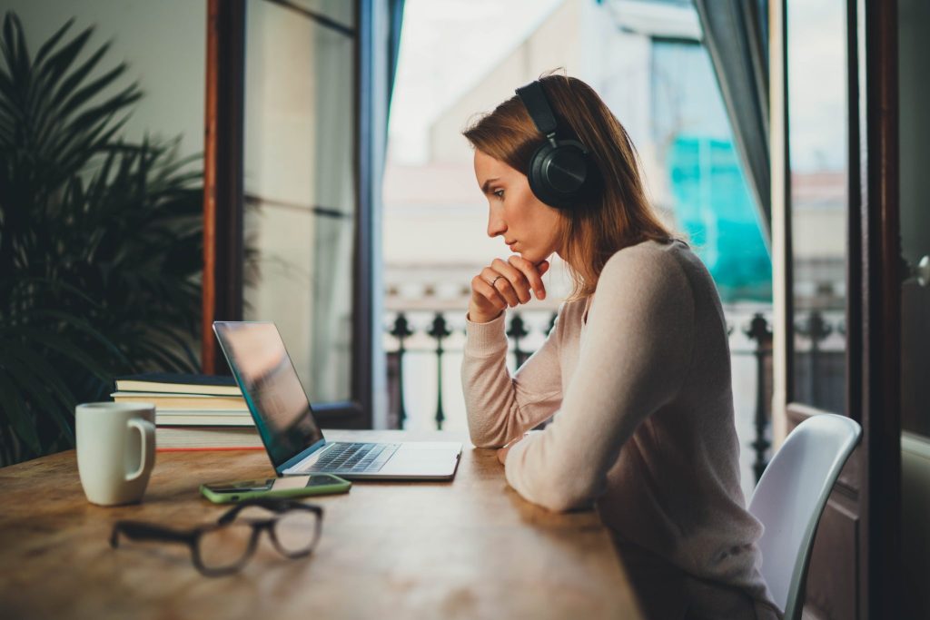 A woman wearing headphones sits at a table with a laptop, staring at the screen, with a cup and eyeglass on the table