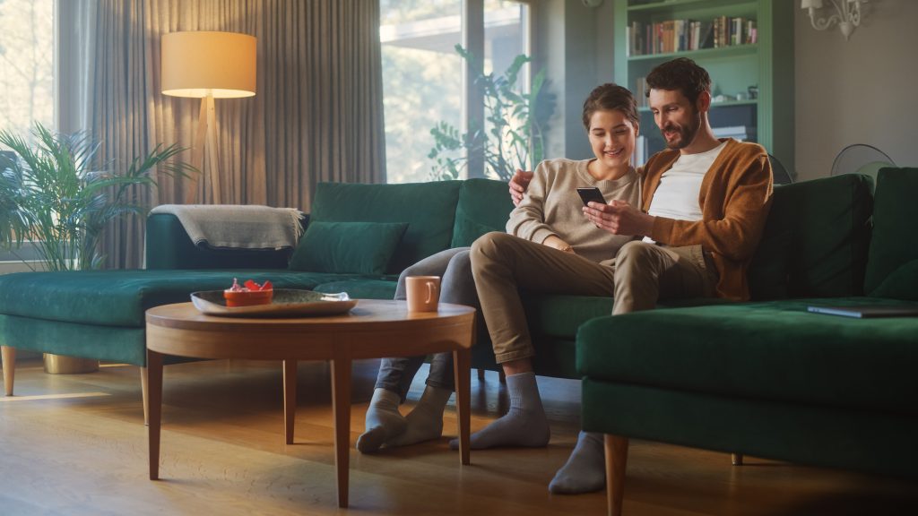 A man and woman sitting on a couch in a living room, watching something on the phone